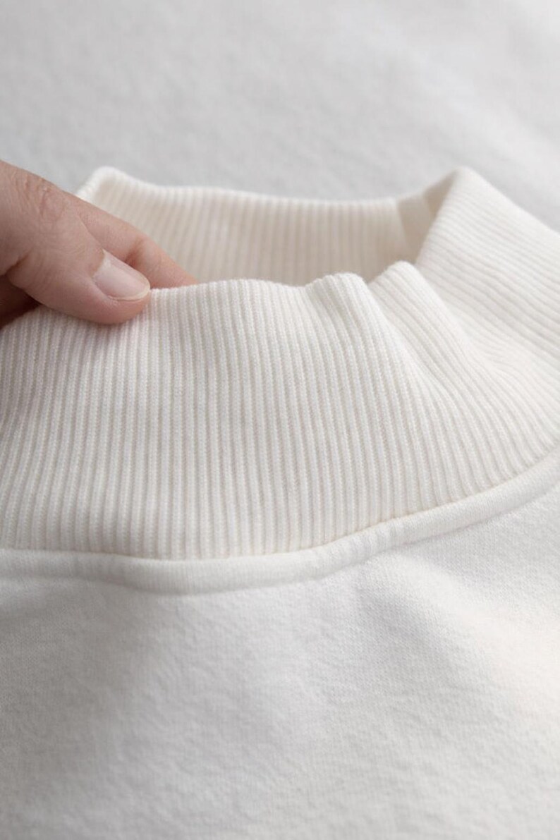 May include: Close-up of a cream-colored sweatshirt collar. The ribbed collar is a lighter shade of cream than the body of the sweatshirt. The fabric appears soft and comfortable. The collar is being touched by a hand.