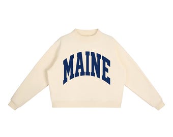 May include: A cream-coloured long-sleeve sweatshirt featuring a mock neck collar and ribbed cuffs. The word MAINE is printed across the chest in a large, arched, dark navy blue collegiate-style font. The garment has a clean, simple design.