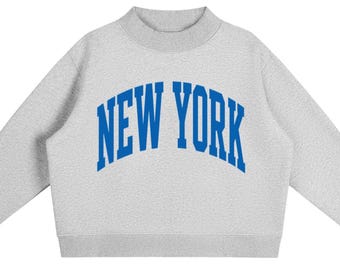 May include: A light grey crewneck sweatshirt featuring a mock neck collar and long sleeves. The front of the garment displays the text NEW YORK printed in a bold, arched, royal blue collegiate-style font across the centre of the chest area.