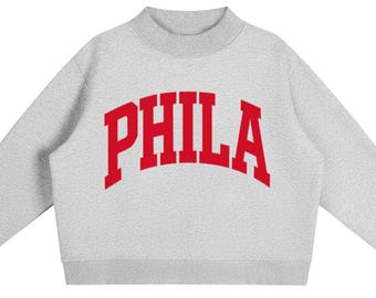 May include: A light grey long-sleeve sweatshirt with a mock neck collar and ribbed cuffs. The front features the word PHILA printed in a bold, arched, red collegiate-style font across the chest. The garment has a textured, fleece-like appearance.