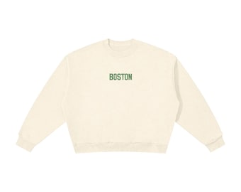 May include: A cream-coloured crewneck sweatshirt featuring the word BOSTON printed in dark green capital letters across the centre of the chest. The garment has long sleeves with ribbed cuffs and a ribbed hemline, providing a simple design.