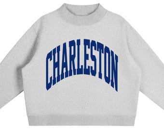 May include: A light grey crewneck sweatshirt featuring a mock neck collar and long sleeves. The word CHARLESTON is printed across the chest in a bold, dark blue, arched collegiate-style font. The garment has ribbed cuffs and a ribbed hem.