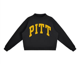 Op de afbeelding: Een zwart sweatshirt met lange mouwen, een opstaande kraag en geribbelde boorden. Het woord PITT is in een groot, gebogen, geel lettertype in collegestijl in het midden op de borst gedrukt, wat een opvallend contrast vormt met de zwarte stof.