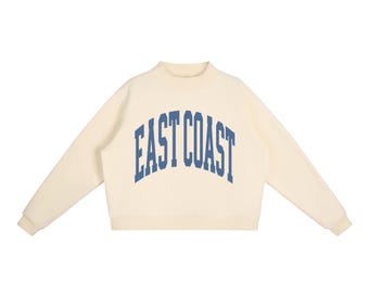 May include: A cream-coloured long-sleeve sweatshirt featuring a mock neck collar and ribbed cuffs. The front of the garment displays the text EAST COAST printed in a bold, arched blue collegiate-style font across the chest area.