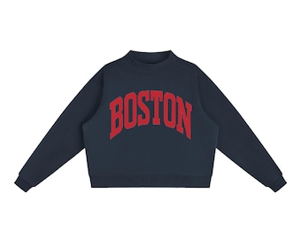 May include: A dark navy blue long-sleeve sweatshirt featuring a mock neck collar and ribbed cuffs. The word BOSTON is printed in a large, arched, red collegiate-style font across the centre of the chest, creating a bold contrast against the dark fabric.