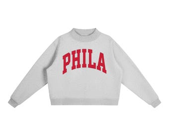 May include: A light gray sweatshirt with the word "PHILA" in red, arched lettering. The sweatshirt has a crew neck and long sleeves with gray cuffs. The garment is made of a soft, textured material.