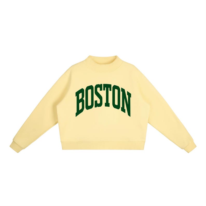 Op de afbeelding: Een lichtgeel sweatshirt met lange mouwen, een opstaande kraag en geribbelde boorden. Het woord BOSTON is in een donkergroen, gebogen lettertype in collegestijl op de borst gedrukt. Het kledingstuk heeft een rechte zoom en lange mouwen.