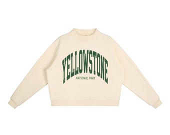 May include: A cream-coloured long-sleeve sweatshirt featuring a mock neck and ribbed cuffs. The front displays the text YELLOWSTONE in a large, arched, distressed dark green font, with the words NATIONAL PARK printed below in a smaller font.