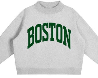 May include: A light grey crewneck sweatshirt featuring the word BOSTON printed in a dark green, arched collegiate-style font across the chest. The garment has long sleeves with ribbed cuffs, a ribbed hem, and a ribbed mock neck collar design.