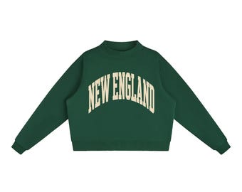 May include: A dark green long-sleeve sweatshirt featuring a mock neck collar and ribbed cuffs. The chest displays the text NEW ENGLAND in a large, arched, cream-coloured collegiate-style font centred across the front of the garment.