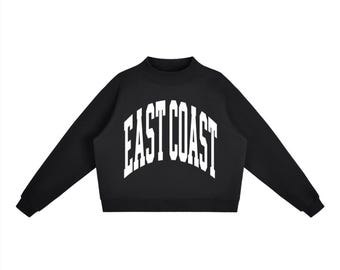 May include: A black long-sleeve sweatshirt featuring a mock neck collar and ribbed cuffs. The front of the garment displays the text "EAST COAST" in a large, white, arched collegiate-style font centred across the chest area of the shirt.
