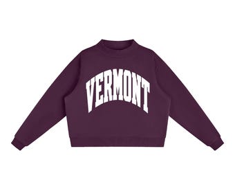 May include: A dark purple long-sleeve sweatshirt featuring a mock neck collar and ribbed cuffs. The word VERMONT is printed in large, white, arched capital letters across the centre of the chest, creating a bold contrast against the fabric.
