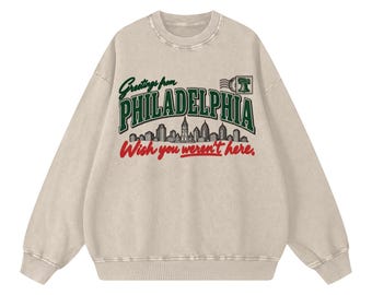 May include: A beige crewneck sweatshirt featuring a graphic print that reads Greetings from PHILADELPHIA in dark green, with a black and white city skyline illustration, a small Liberty Bell icon, and the red text Wish you weren't here.