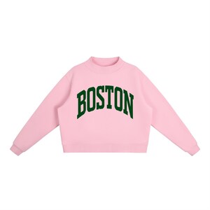Op de afbeelding: Een lichtroze sweatshirt met lange mouwen, een opstaande kraag en geribbelde boorden. Het woord BOSTON is in een donkergroen, gebogen lettertype in collegestijl in het midden op de borst gedrukt. Het kledingstuk heeft een rechte zoom.