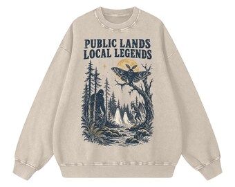 May include: A beige crewneck sweatshirt featuring the text "PUBLIC LANDS LOCAL LEGENDS" above a dark blue graphic illustration of a forest scene with a large moth, a creature resembling Bigfoot, two ghosts, and a small dragon near a stream.