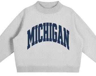 May include: A light grey long-sleeve sweatshirt featuring a mock neck collar and ribbed cuffs. The word MICHIGAN is printed in large, dark navy blue capital letters across the chest in a bold, arched collegiate-style font on the front centre.