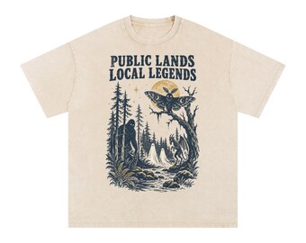 May include: A beige short-sleeve t-shirt featuring a dark blue graphic print with the text "PUBLIC LANDS LOCAL LEGENDS". The illustration depicts a forest scene with a moth, a large creature, two ghosts, a dragon, and a full moon.
