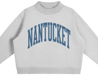 May include: A light grey long-sleeve sweatshirt with a mock neck collar and ribbed cuffs. The front features the word NANTUCKET printed in a large, arched, dark blue collegiate-style font across the chest area of the heathered grey garment.