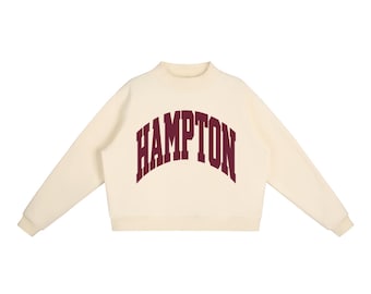 May include: A cream-coloured long-sleeve sweatshirt featuring a mock neck collar and ribbed cuffs. The word HAMPTON is printed in a large, arched, dark maroon collegiate-style font across the centre of the chest area of the garment.