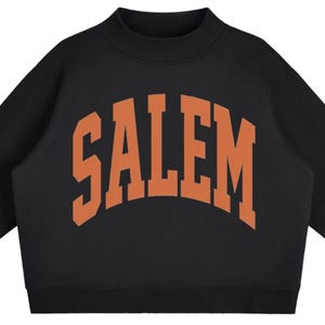 May include: A black long-sleeve sweatshirt featuring a mock neck collar and ribbed cuffs. The word SALEM is printed across the chest in a large, arched, orange collegiate-style font. The garment has a solid black base color with a contrasting orange graphic.