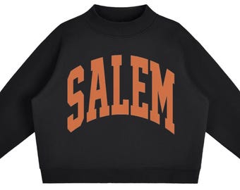 May include: A black long-sleeve sweatshirt featuring a mock neck collar and ribbed cuffs. The word SALEM is printed across the chest in a large, arched, orange collegiate-style font. The garment has a solid black base colour with a contrasting orange graphic.