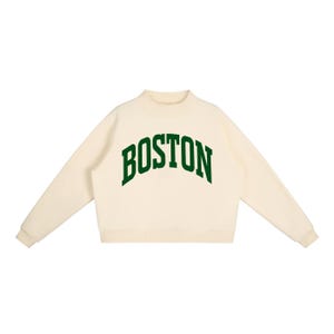 May include: Cream-colored sweatshirt with the word "BOSTON" arched across the chest in bold, green letters. The sweatshirt has a mock neck and long sleeves. A casual, comfortable top.