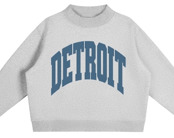 May include: A light grey long-sleeve sweatshirt featuring a mock neck collar and ribbed cuffs. The word DETROIT is printed across the chest in a large, arched, dark blue collegiate-style font. The garment has a simple, clean design.