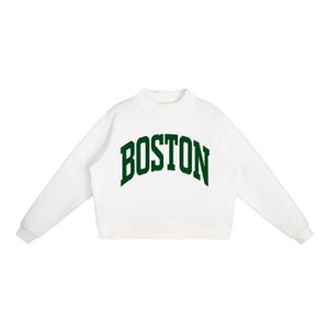 Op de afbeelding: Een wit sweatshirt met lange mouwen, een opstaande kraag en geribbelde boorden. Het woord BOSTON staat in een grote, donkergroene, gebogen lettertype in collegestijl op de borst gedrukt. Het kledingstuk heeft een strak en eenvoudig ontwerp.