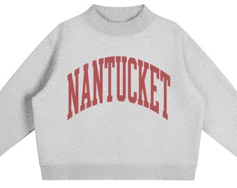 May include: A light grey long-sleeve sweatshirt with a mock neck collar. The front features the word NANTUCKET printed in a bold, arched, red serif font. The garment has ribbed cuffs at the sleeves and a ribbed hemline, providing a structured fit.