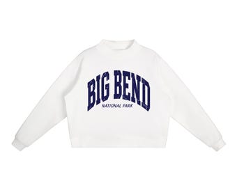 May include: A white long-sleeve sweatshirt featuring a mock neck collar and ribbed cuffs. The chest displays the text BIG BEND in a large, dark blue, distressed-style arched font, with the words NATIONAL PARK printed in a smaller blue font below.