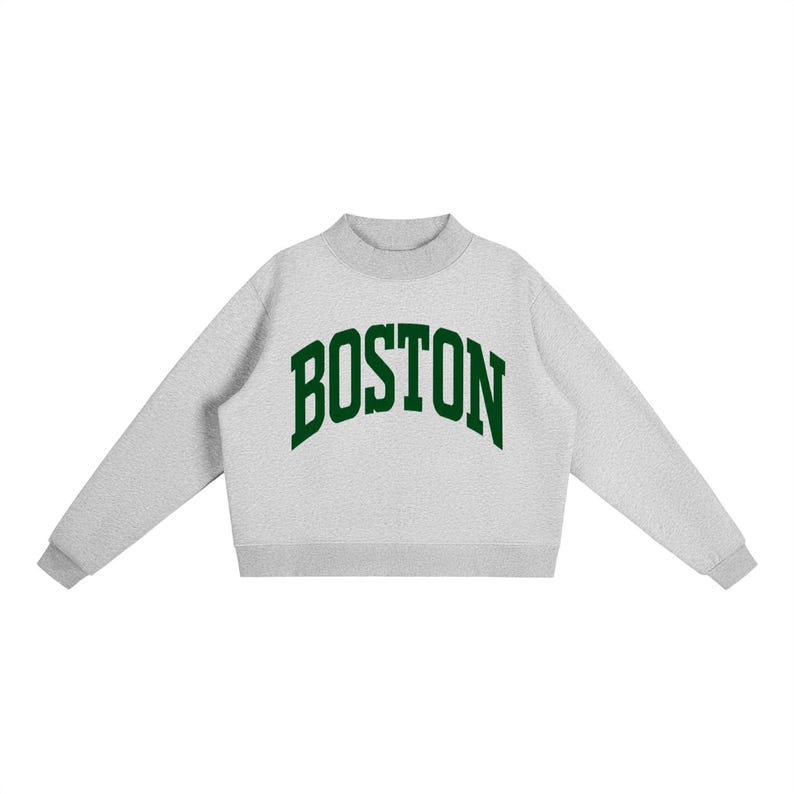 Op de afbeelding: Een lichtgrijs sweatshirt met ronde hals, lange mouwen en een geribbelde kraag. Het woord BOSTON staat op de borst gedrukt in een donkergroen, gebogen lettertype in collegestijl. Het kledingstuk heeft een rechte zoom en een textuur.