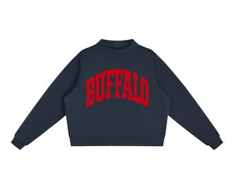 May include: A dark navy blue long-sleeve sweatshirt featuring a mock neck collar and ribbed cuffs. The word BUFFALO is printed across the chest in a bold, arched, red collegiate-style font. The garment has a clean, solid-coloured design.