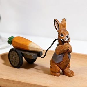May include: A wooden carved figurine of a brown rabbit pulling a cart with a large orange carrot. The rabbit wears a gray vest and holds the cart's black pull cord. The carrot has a green leafy top and is resting on a dark gray cart.
