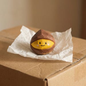 Könnte beinhalten: Handgeschnitzte Holz-Kastanienfigur mit einem lächelnden Gesicht, bemalt in Gelb und Braun. Die Kastanie liegt auf einem Stück weißem Papier, das auf einem Karton platziert ist. Die Figur hat ein skurriles, cartoonartiges Design.