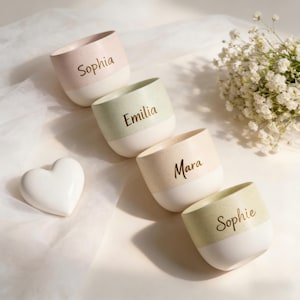 May include: Four small, speckled ceramic cups in pastel shades, each with a white base and a name printed on the side: Sophia, Emilia, Mara, and Sophie. A white heart-shaped object and a sprig of white flowers are also present.
