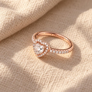 May include: A rose-tone metal ring featuring a central heart-shaped clear stone surrounded by a halo of smaller clear stones. The band is also set with small clear stones and has the word PANDORA engraved on the inner surface.