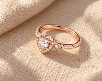 May include: A rose-tone metal ring featuring a central heart-shaped clear stone surrounded by a halo of smaller clear stones. The band is also set with small clear stones and has the word PANDORA engraved on the inner surface.
