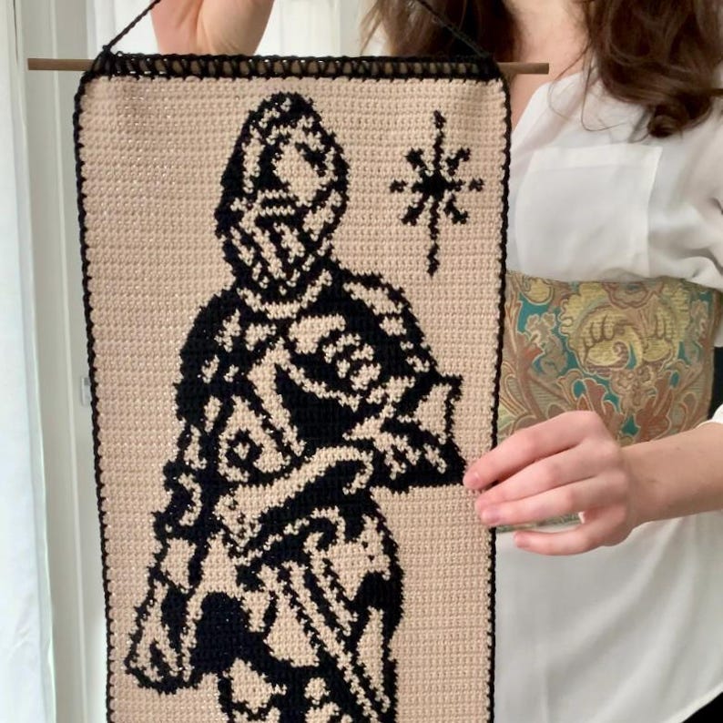 May include: A beige and black crochet wall hanging featuring a stylized figure and a starburst design. The artwork is rectangular, with a black border and a wooden dowel for hanging. The figure is depicted in black against a beige background.