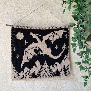 May include: A black and beige crochet wall hanging featuring a dragon silhouette against a night sky with a moon and stars. The bottom of the piece depicts a forest. The wall hanging is suspended from a wooden dowel.