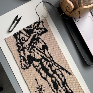 May include: A partially completed crochet project featuring a beige background with a black abstract design. Black scissors and a wooden yarn bowl with yarn are visible. The project is on a white surface.