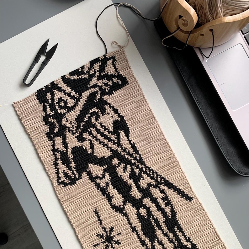May include: A partially completed crochet project featuring a beige background with a black abstract design. Black scissors and a wooden yarn bowl with yarn are visible. The project is on a white surface.