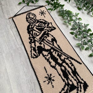 May include: A beige and black crochet wall hanging featuring a stylized figure with a starburst design. The rectangular piece is suspended from a wooden dowel with a black cord. The figure is black against a beige background.