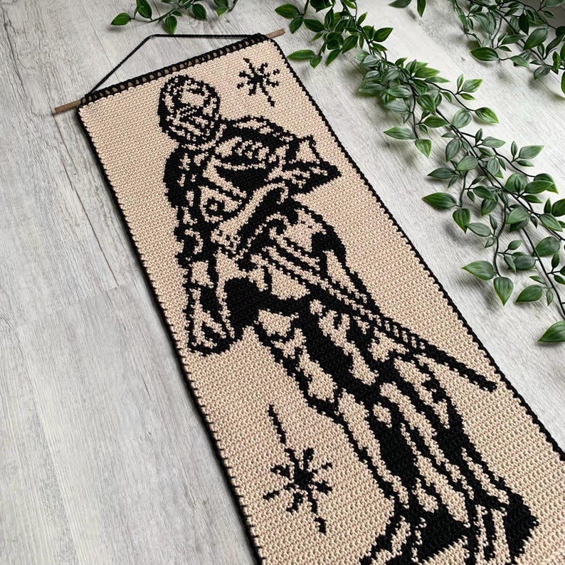 May include: A beige and black crochet wall hanging featuring a stylized figure with a starburst design. The rectangular piece is suspended from a wooden dowel with a black cord. The figure is black against a beige background.