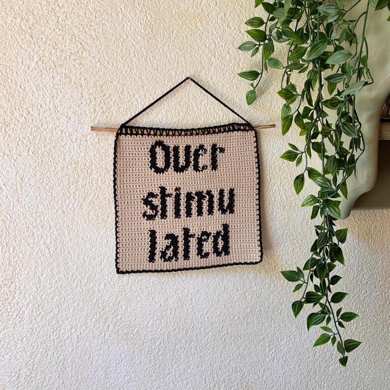 Digital crochet pattern for a wall tapestry featuring the word "Overstimulated" in bold black gothic-style lettering on a beige background. The pattern includes instructions to create the tapestry, which is bordered with black crochet edging. This listing is for the pattern only, not a finished item.