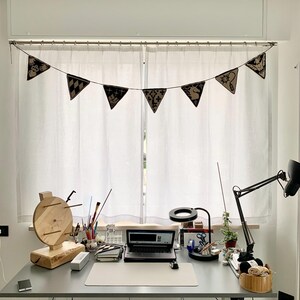 May include: A home office workspace with a gray desk, a laptop, and a wooden yarn swift. A banner of black and tan pennants hangs above the window. A black desk lamp and a magnifying glass are also on the desk.