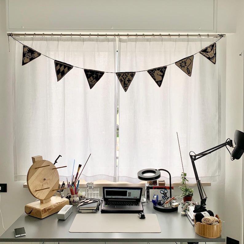 May include: A home office workspace with a gray desk, a laptop, and a wooden yarn swift. A banner of black and tan pennants hangs above the window. A black desk lamp and a magnifying glass are also on the desk.