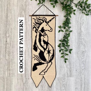 Puede incluir: Un tapiz de ganchillo rectangular con la silueta de un unicornio negro sobre un fondo crema. El diseño incluye un detalle de estrella en el flanco del unicornio. La pieza cuelga de un palo de madera con un cordel negro y tiene dos extremos en punta.