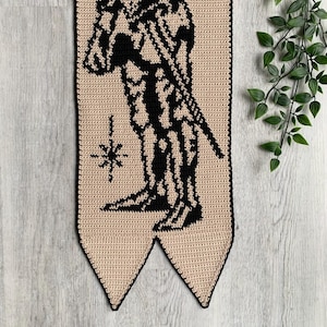 May include: Crocheted wall hanging featuring a black silhouette of a figure holding a sword, set against a beige background. A star-like design is below the figure. The banner has a black border and a pointed bottom.