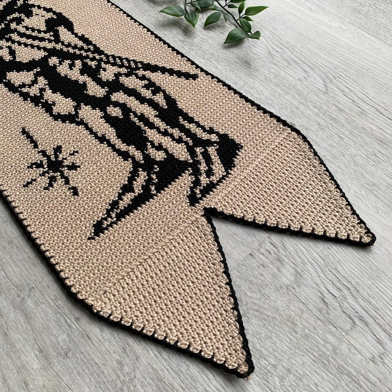 May include: A beige and black crochet banner with a geometric design. The banner features a black figure and star on a beige background, with a black border. The banner is made of yarn and has a unique shape.