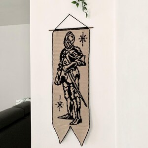 May include: A crochet wall hanging featuring a black silhouette of a knight in armor holding a sword. The knight is set against a tan background with black trim and star-like accents. The banner is suspended from a wooden dowel.